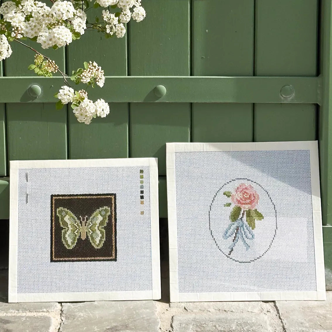 Two framed needlework or embroidery panels leaning against a green wooden door outdoors. One panel shows a butterfly design, and the other shows a pink rose in an oval frame.