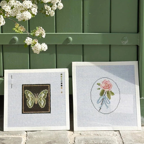 Two framed needlework or embroidery panels leaning against a green wooden door outdoors. One panel shows a butterfly design, and the other shows a pink rose in an oval frame.